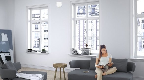 Ein hoher, heller Raum mit einem grauen Sofa. Auf dem Sofa sitzt eine Frau die eine Zeitschrift  liest.