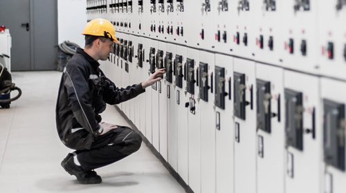 Ein Elektroniker der vor einem Schaltanlagenbau hockt