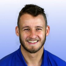 Freundliches Porträt eines jungen Mannes in blauer Kleidung Ein junger Mann mit einem breiten Lächeln trägt ein blaues Hemd und hat einen kurzen Bart, vor einem sanften Hintergrund.